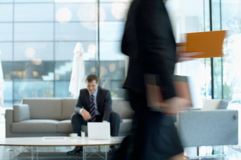 Businessman sitting in modern office lounge using a laptop while colleague walks past, creating dynamic blur.